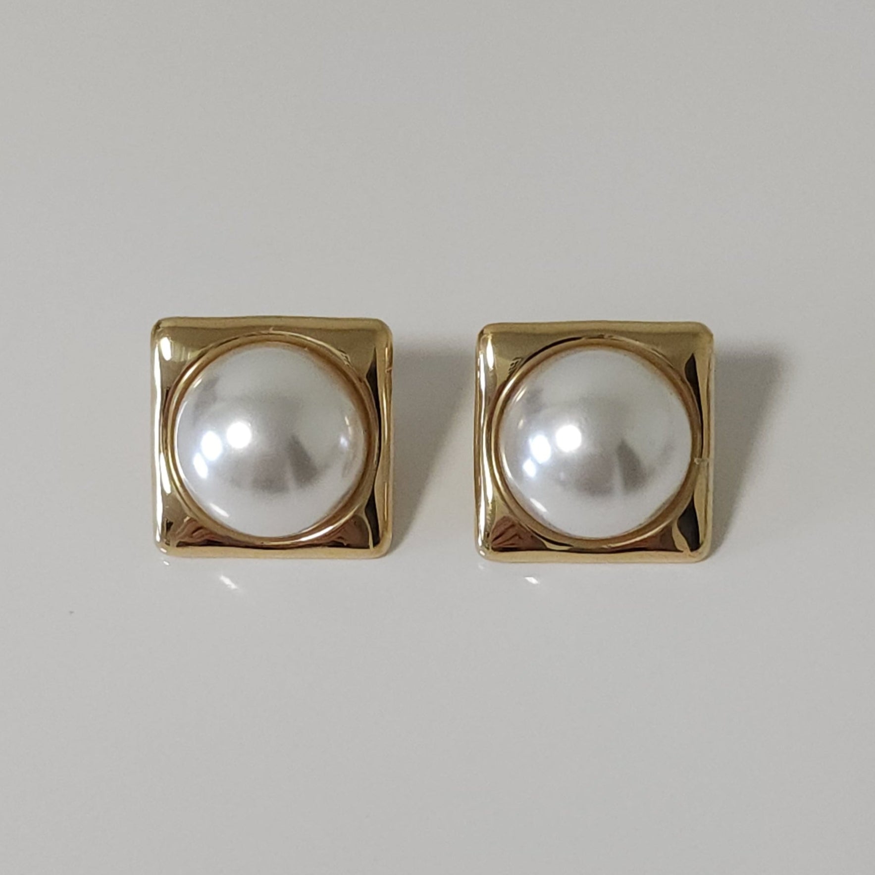 Pair of square gold earrings with pearl-like centers on a light gray background