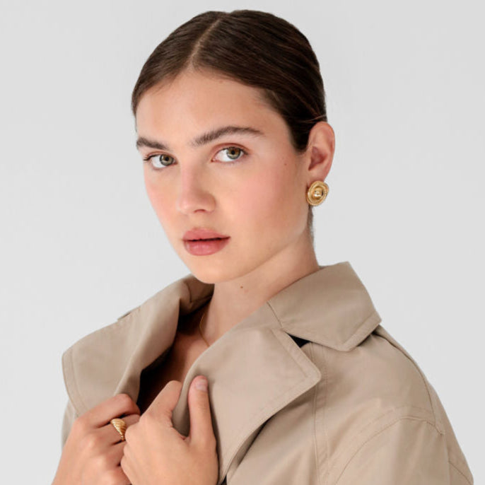 Woman wearing gold statement earrings and a beige trench coat.