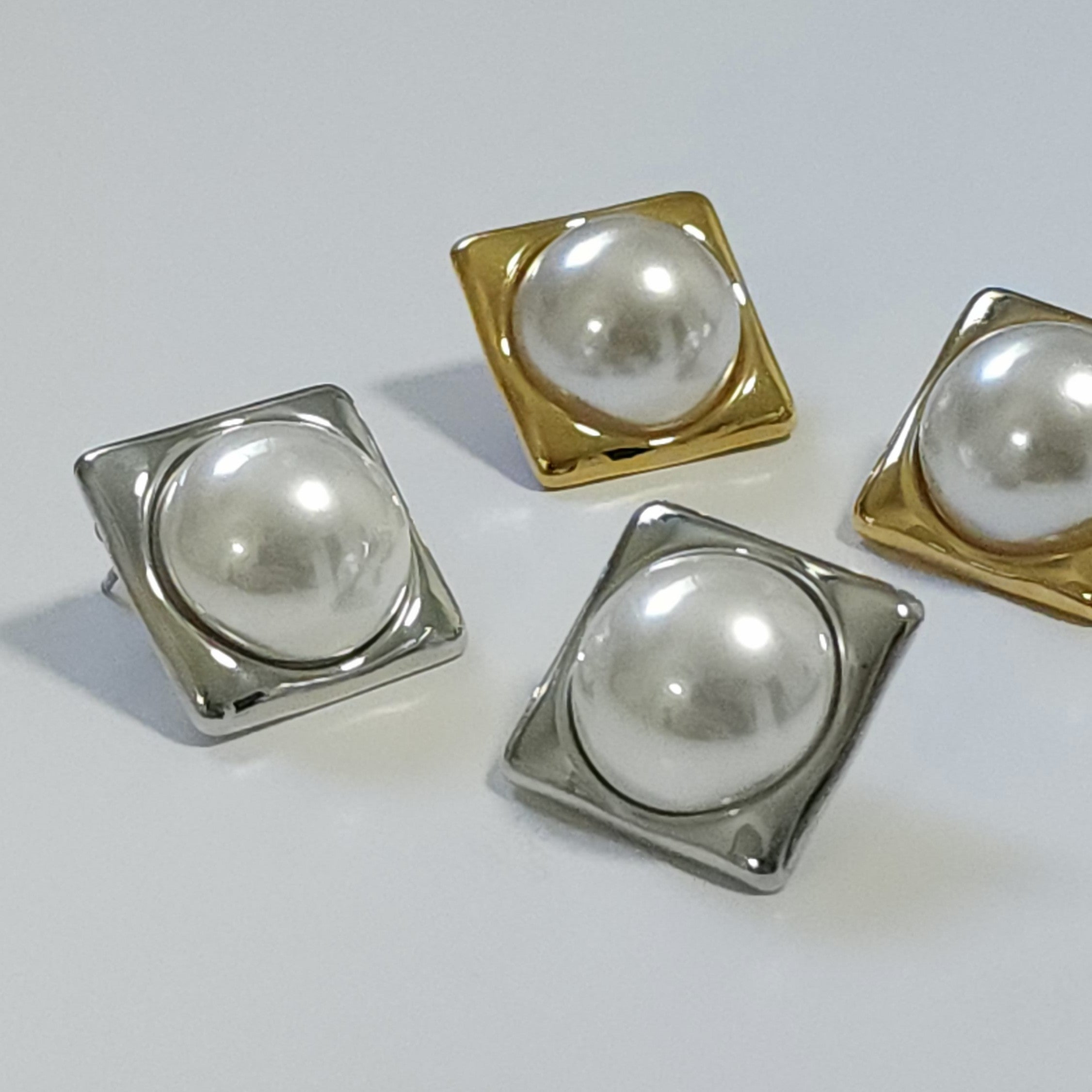 Four pearl earrings with square metal bases in gold and silver on a light gray background