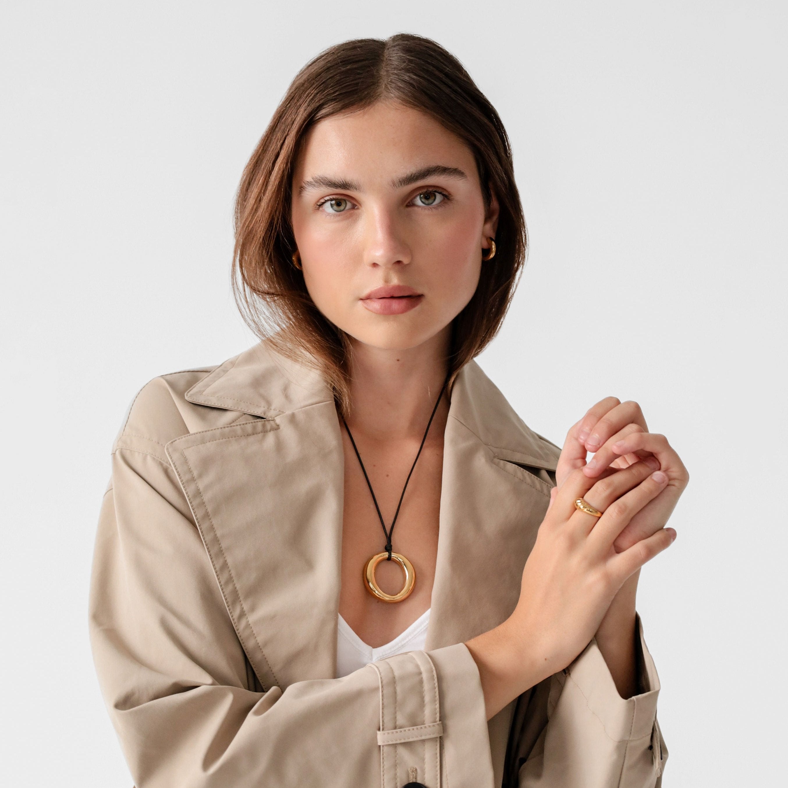 Woman wearing gold pendant necklace with a beige trench coat against a light background.