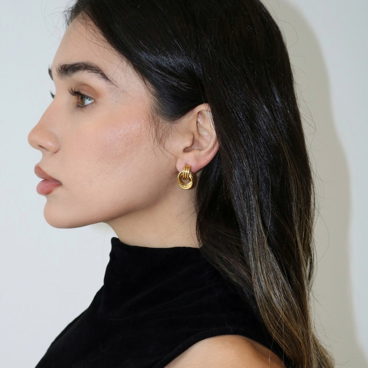 Woman wearing gold stud earrings in a black tank top against a light background.