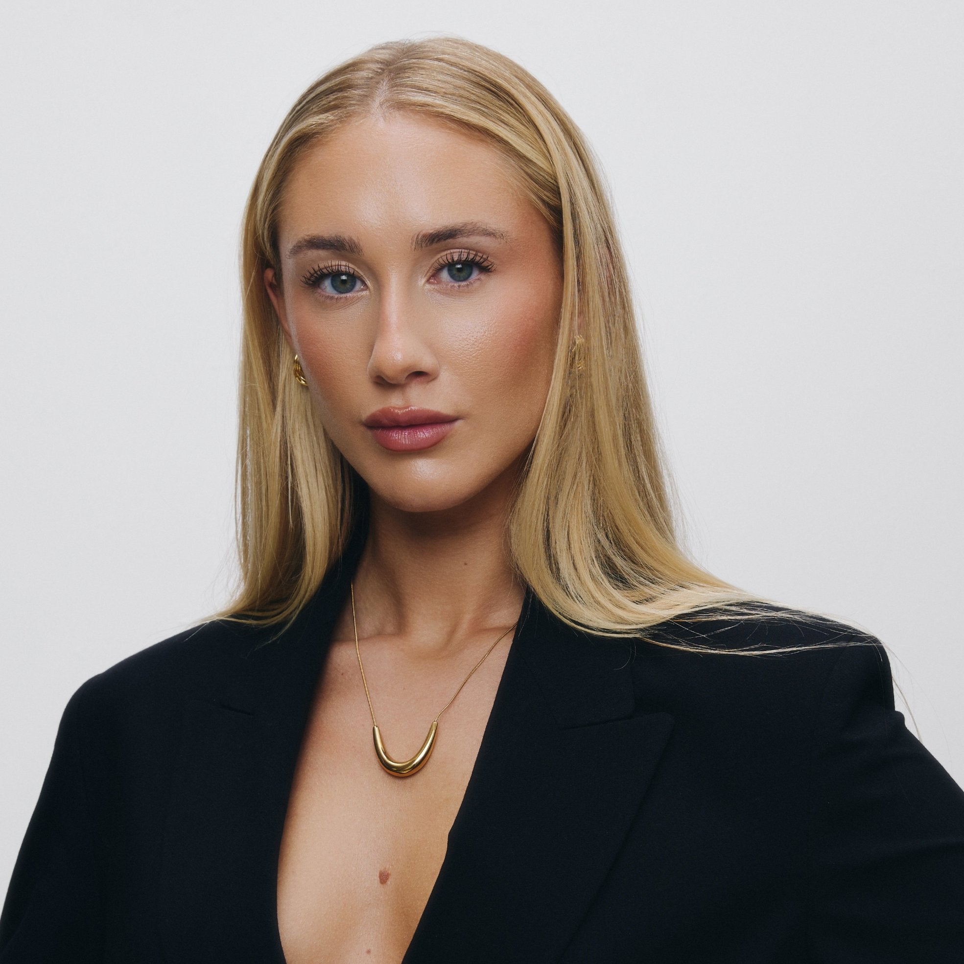 Woman wearing gold pendant necklace and a black blazer against a light background.