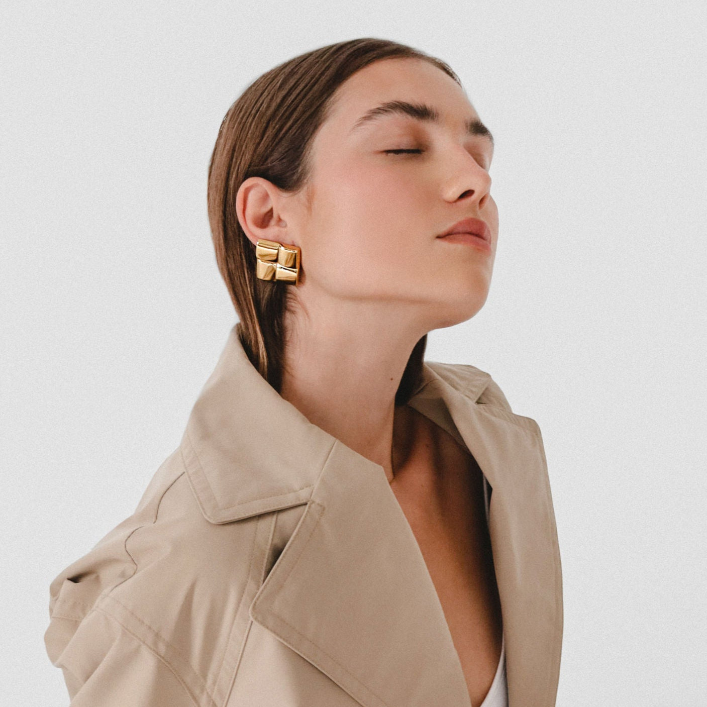 Woman wearing gold earrings with a beige coat on a light background