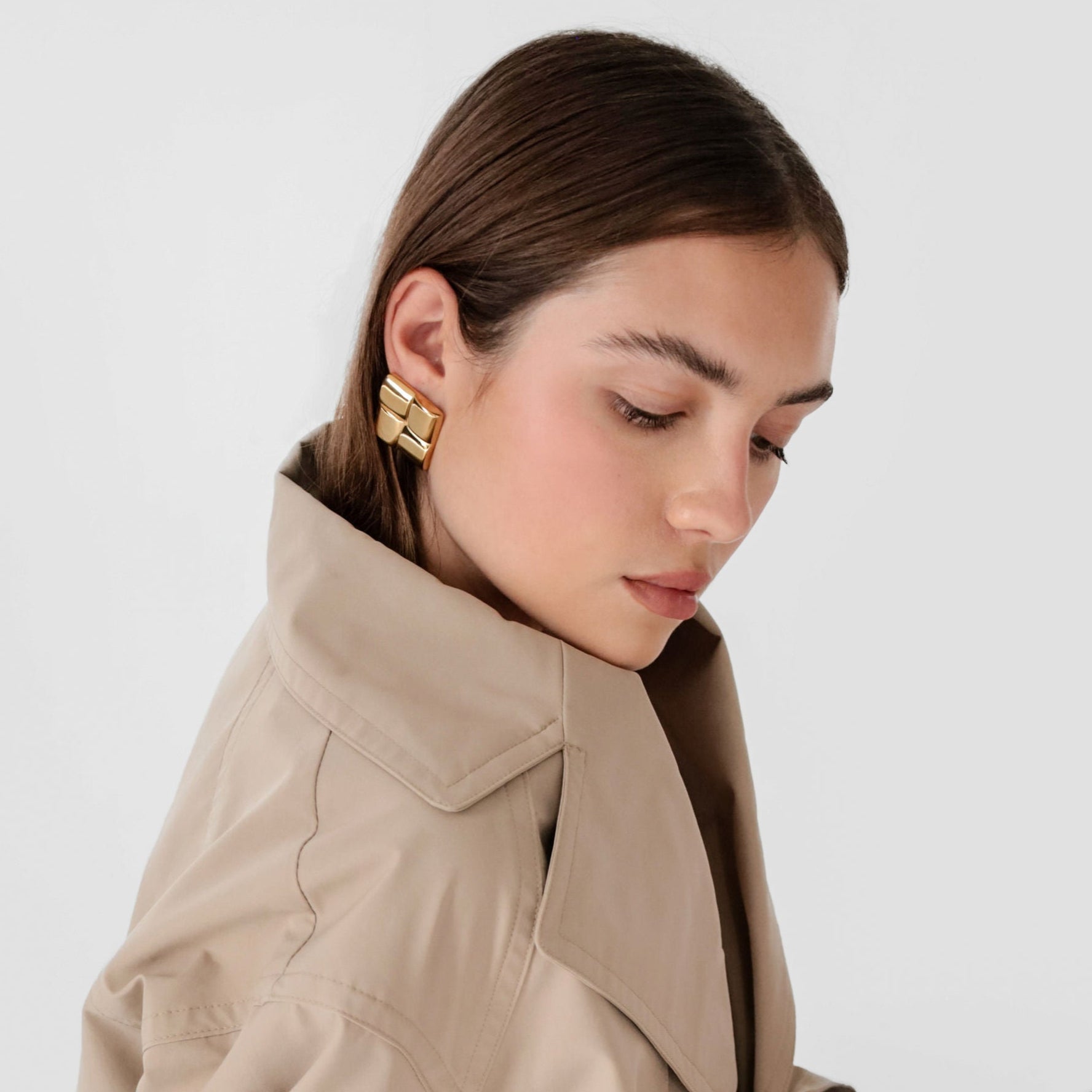 Woman wearing gold square statement earrings and a beige trench coat.