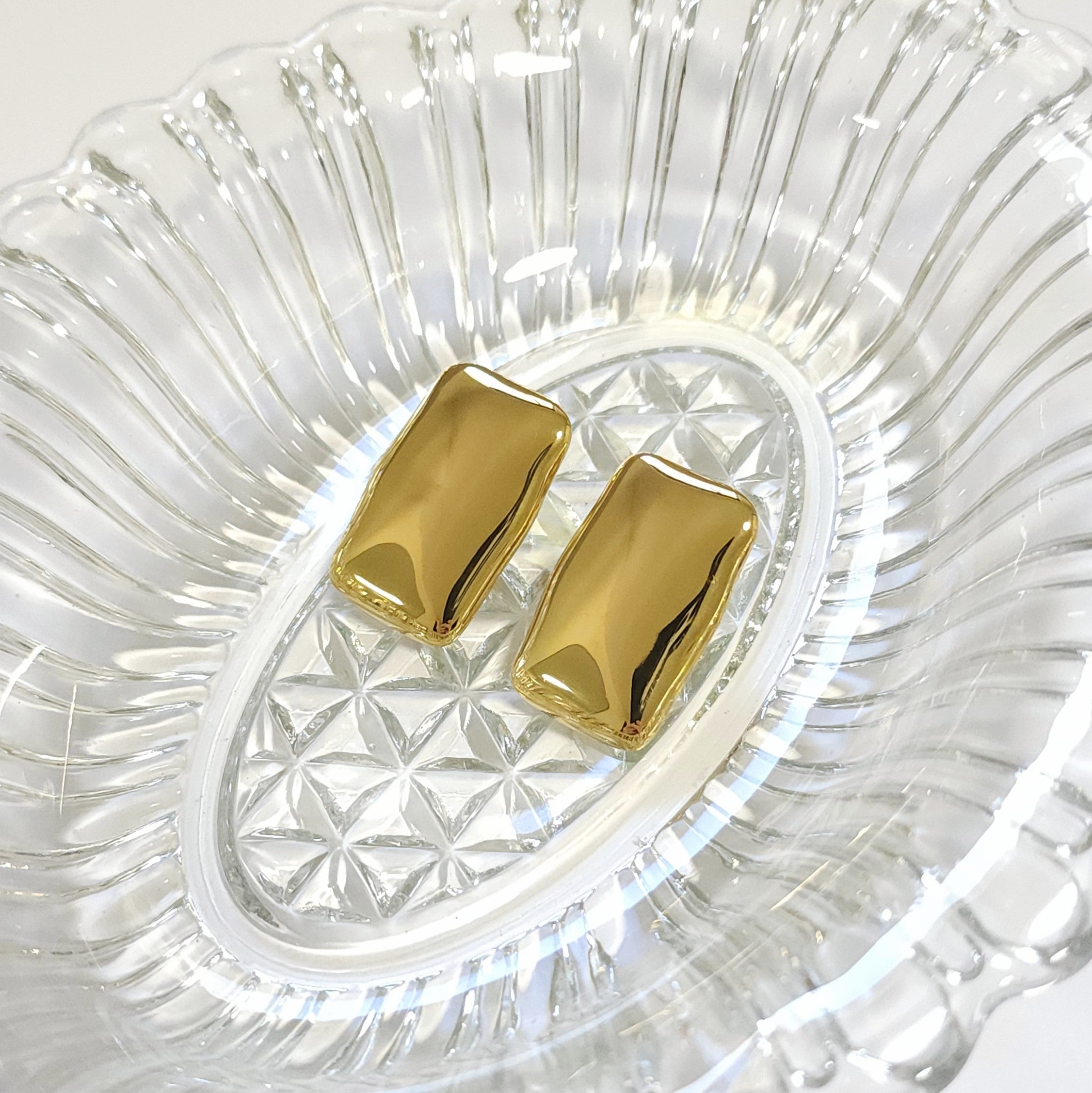 Gold mirrored statement earrings on a clear glass dish.