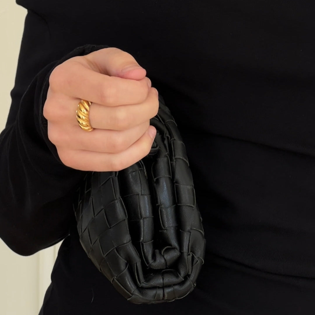 Person holding a black woven clutch with a gold ring on a neutral background